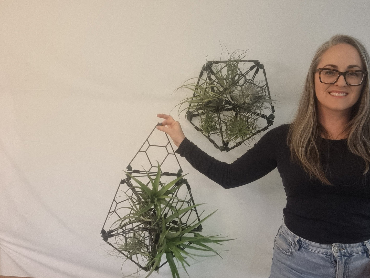 A woman wearing glasses and a black long-sleeve top stands next to a white wall, holding up a geometric hanging planter with green air plants, with a second matching planter hanging on the wall behind her.