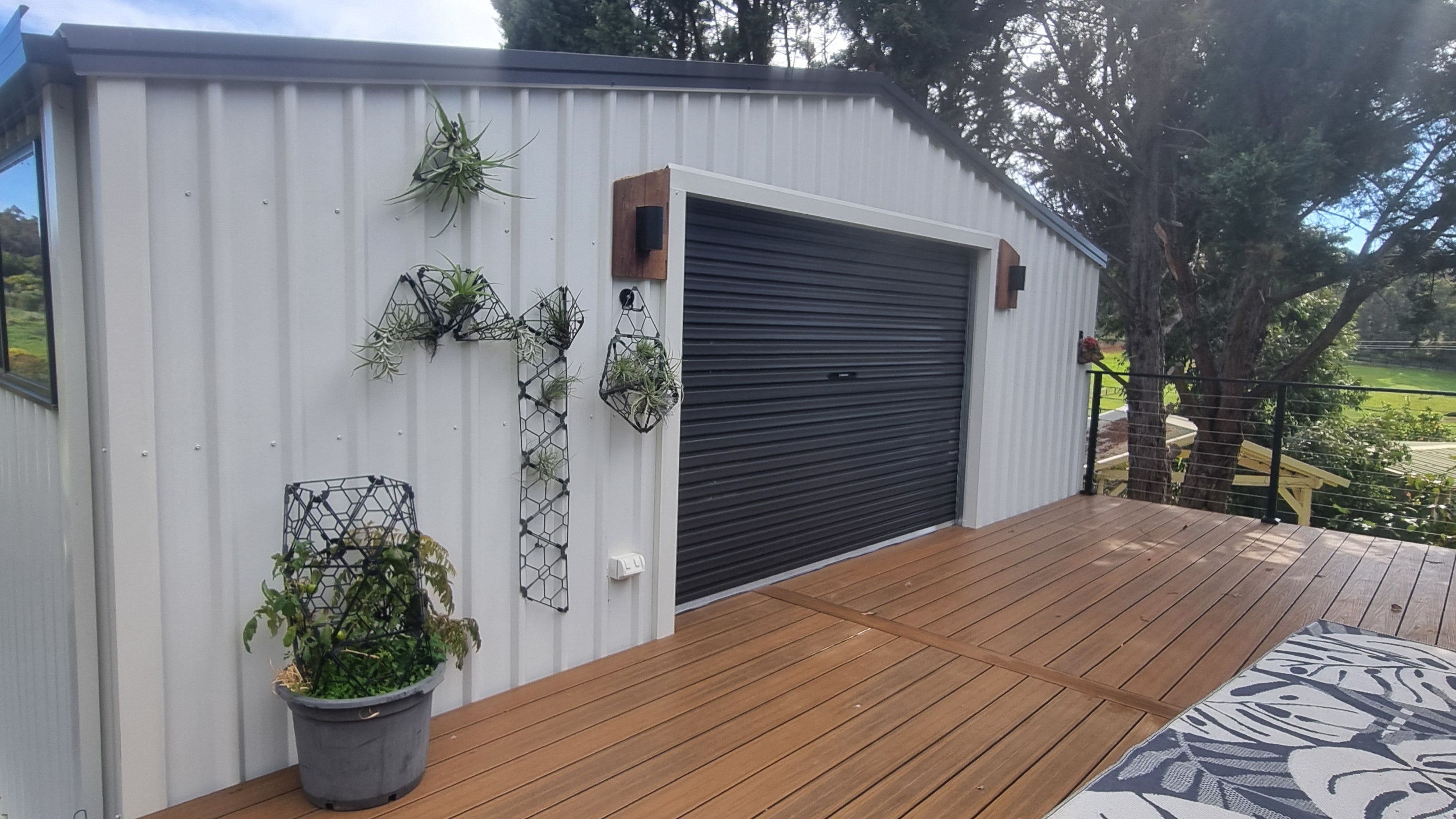 A white metal garage shed with a dark gray roller door, featuring a wooden deck, potted plants, and a scenic outdoor background with trees and grass.